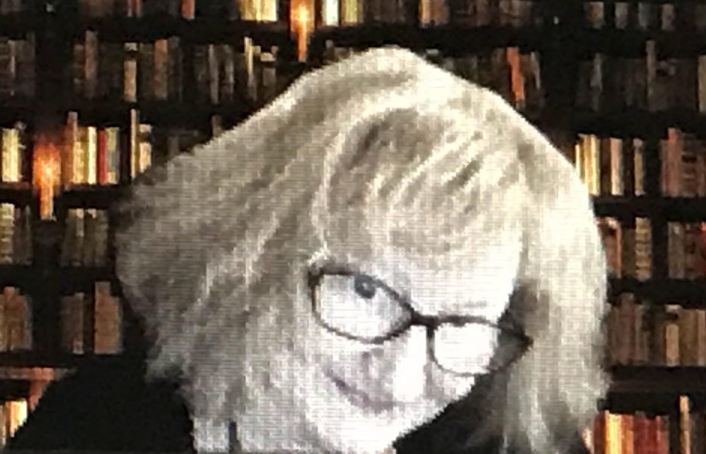 Pixellated face of a woman with blonde hair wearing glasses, smiling at the camera in front of a bookshelf.